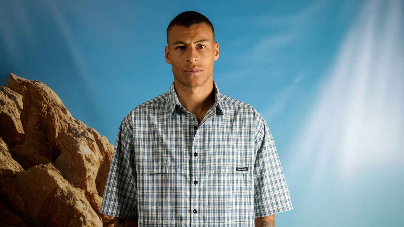St Goliath Clothing. Man wearing checkered shirt in front of rocky backdrop.